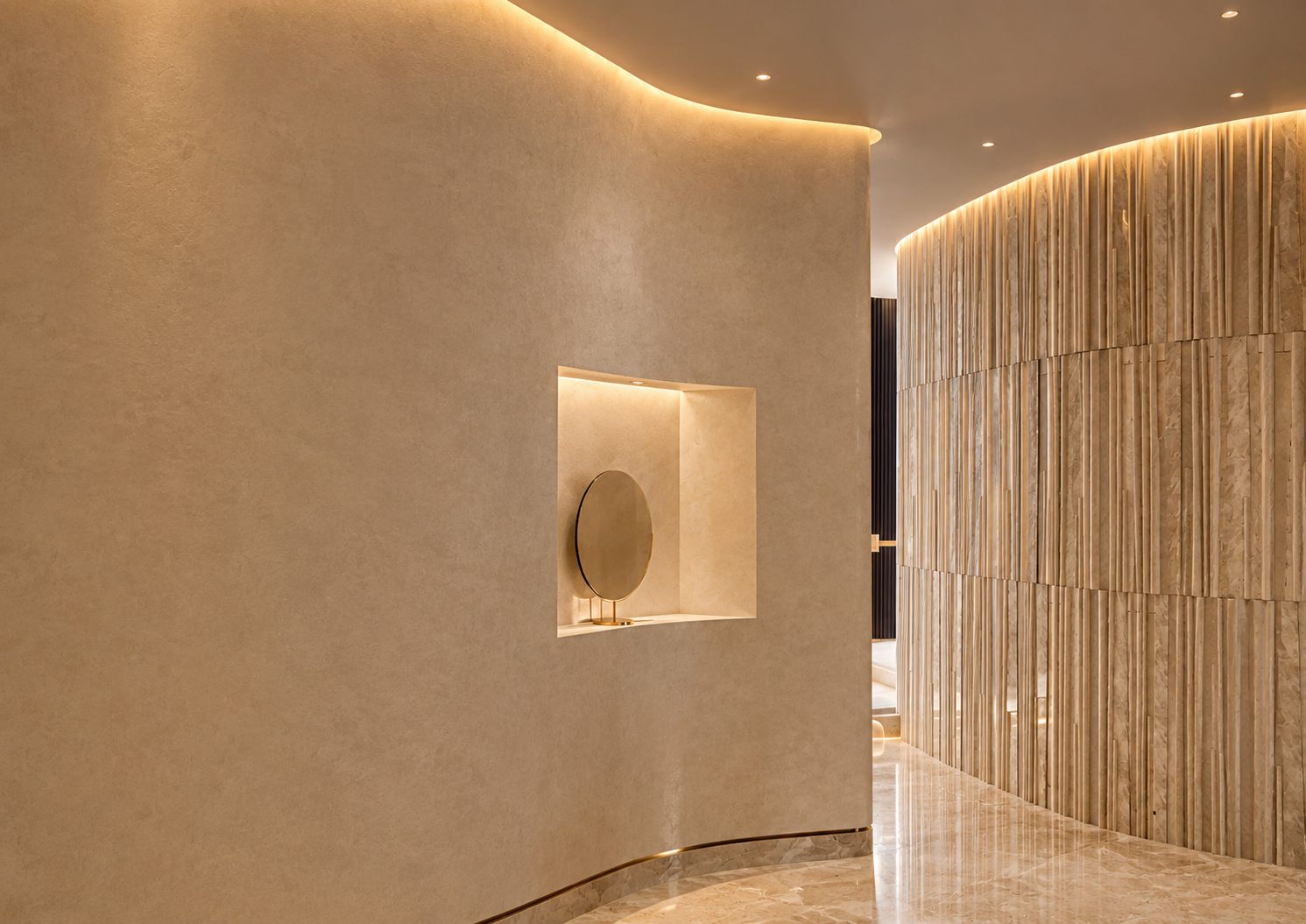 A curved plaster wall with a lit niche holding a round mirror, beside a fluted stone screen wall.